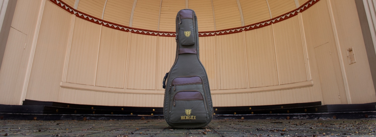 HÉRGÉT guitar case standing upright on a paved surface in front of a curved wooden pavilion with light paneling and decorative red-and-white border.