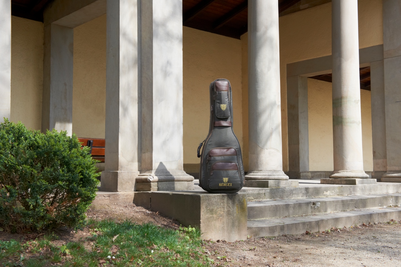 HÉRGÉT guitar case resting on stone steps between tall classical pillars, with greenery and pavilion architecture in the background.