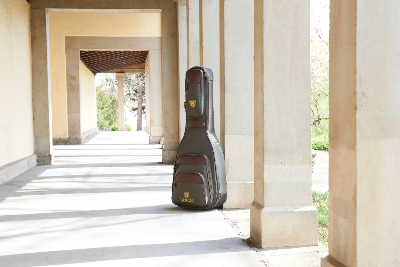 HÉRGÉT guitar case positioned between large stone pillars in a classical colonnade walkway, with sunlight casting long shadows into the distance.
