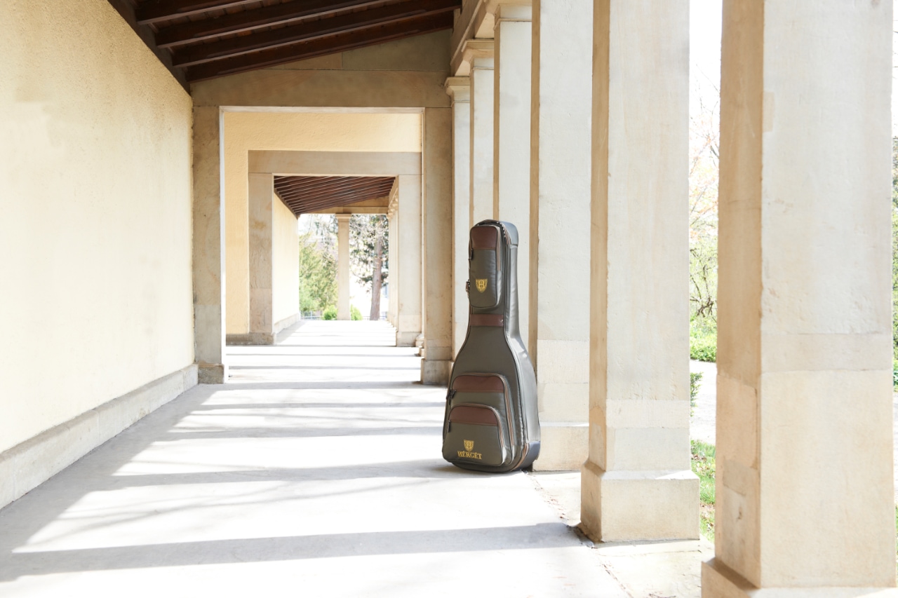 HÉRGÉT guitar case leaning against a stone pillar in a long sunlit colonnade with repeating arches and strong shadows on the walkway.